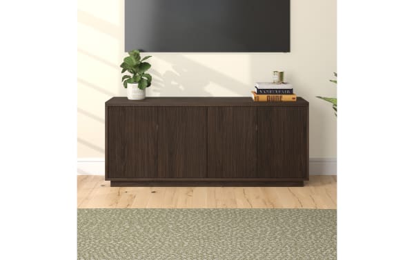 Dark brown wooden sideboard with vertical grooves, four doors, placed on light hardwood floor with green patterned rug, styled with a potted plant and stacked books.