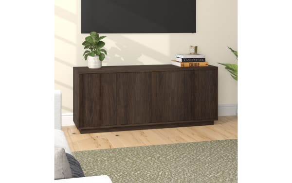Dark brown wooden sideboard with vertical grooves, featuring three doors, placed in a modern living room with light wood flooring and decorative plants.
