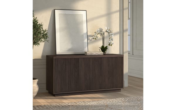 Dark brown wooden sideboard with vertical grooves, topped with a white textured framed artwork, a book, and a white orchid plant, in a sunlit room.