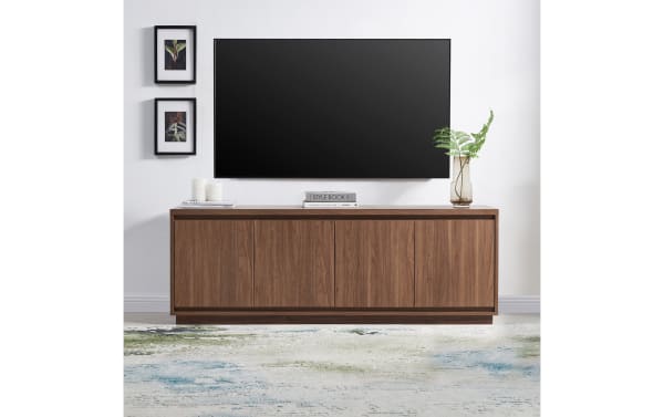 Modern wooden TV console with walnut finish, four cabinet doors, minimalist design, paired with wall-mounted flat-screen TV and decorative plants.