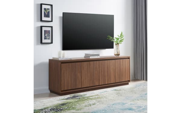 Modern wooden TV console with walnut finish, featuring four cabinet doors, styled with books, candles, and a glass vase with green fern, under a wall-mounted flat-screen TV.