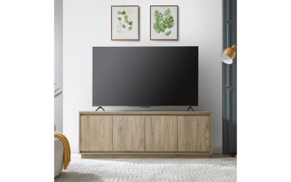 Modern living room setup with a large flat-screen TV on a light wood grain TV stand featuring four cabinet doors, complemented by botanical wall art and a gold floor lamp.