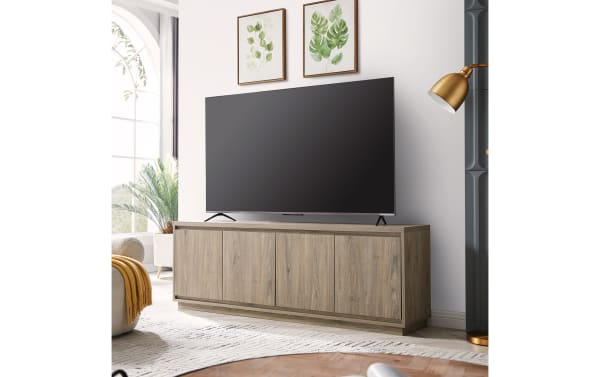 Modern wooden TV stand with a natural oak finish, featuring four cabinet doors and a large flat-screen TV on top, styled in a bright living room.