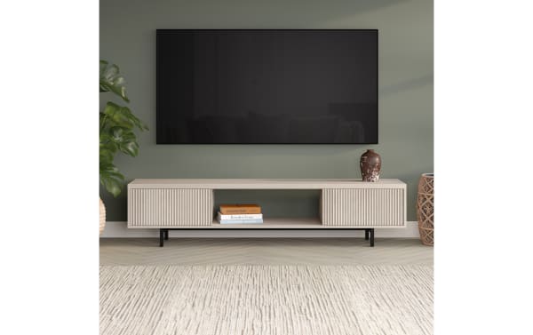 Modern beige wood TV stand with vertical grooves, black metal legs, open shelf with books, and a large flat-screen TV mounted on a muted green wall.