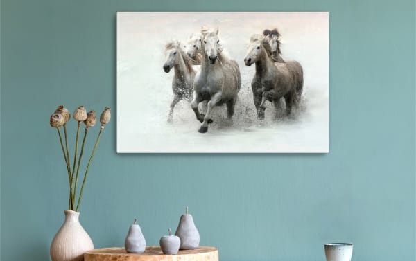 Five white horses running through water, displayed as a wall art canvas in a modern room with blue-green walls and decorative vases.