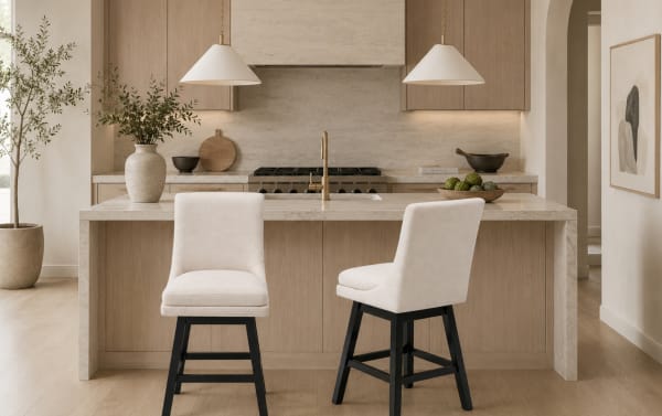 Modern kitchen interior with beige marble countertops, light wood cabinetry, two cream upholstered bar stools with black legs, pendant lighting, and decorative plants.