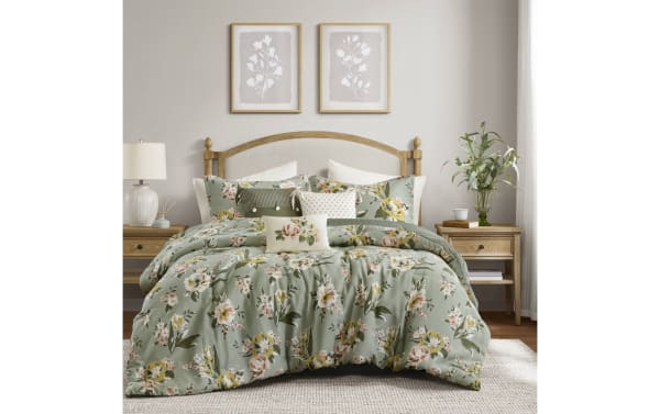 Bed with floral green comforter set, wooden headboard, matching pillows, light wood nightstands with lamp and plant, neutral bedroom decor.