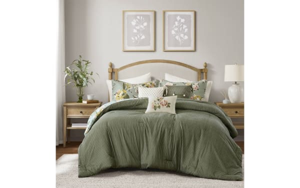 King-size bed with green striped comforter, floral accent pillows, wooden headboard, matching wooden nightstands with lamp and plant, neutral bedroom decor.