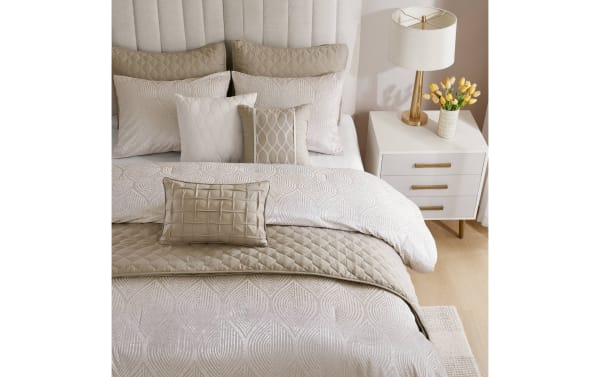 Luxurious beige and cream quilted bedding set with textured decorative pillows on a tufted headboard bed, paired with a white nightstand, gold lamp, and yellow tulip flowers.