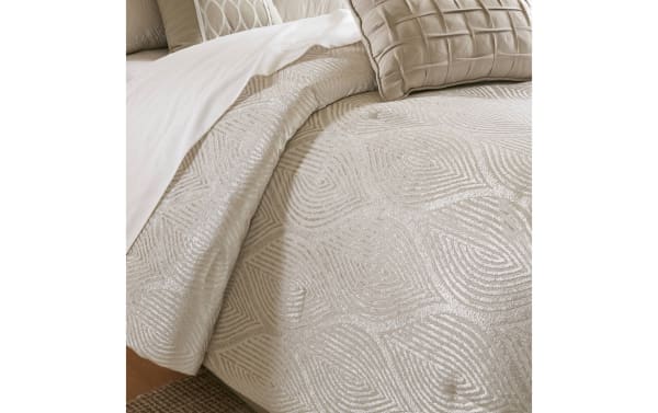 Beige textured comforter with geometric swirl patterns, paired with matching beige decorative pillows and white sheets on a bed.