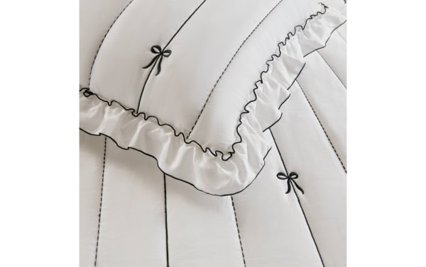 White quilted bedding set with black embroidered bows, vertical black stitching, and ruffled edges, showcasing elegant contrast detailing.