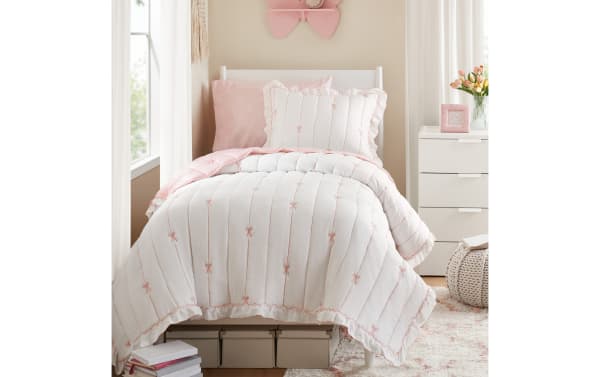 White and pink twin bed set with ruffled edges and embroidered bows, featuring a comforter, pillow shams, and floral accent pillow in a cozy bedroom setting.