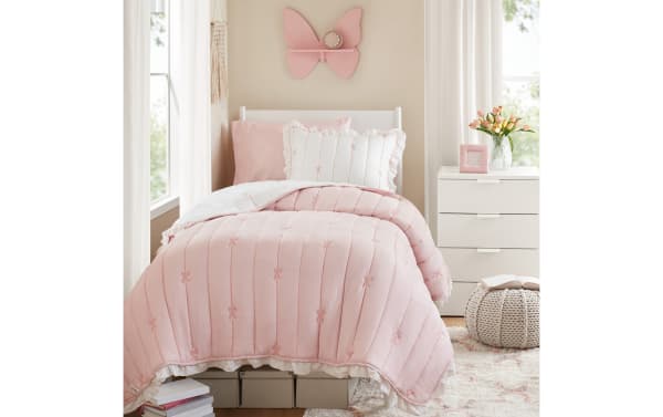 Twin bed with a pink quilted comforter featuring embroidered bows, white ruffled edges, matching pillows, butterfly wall clock, and white nightstand with flowers.
