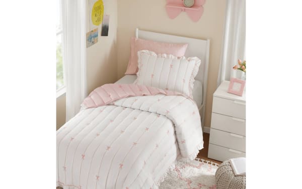 Twin bed with white and pink quilted comforter featuring embroidered bows, matching pillow shams, pink accent pillow, and white wooden headboard in a cozy bedroom setting.