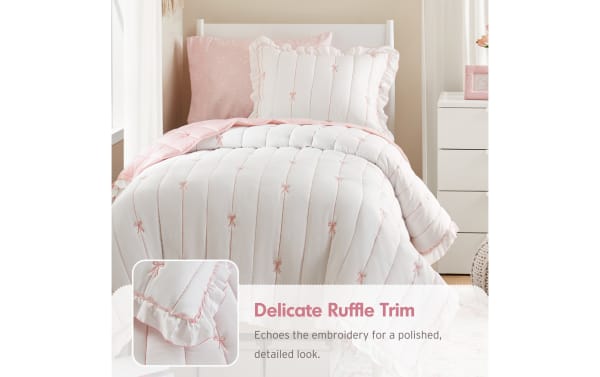 White and pink comforter set with delicate ruffle trim and embroidered bows on a twin bed, highlighting polished, detailed bedding design. Text reads: "Delicate Ruffle Trim. Echoes the embroidery for a polished, detailed look."