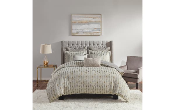 Queen-size bed with tufted gray upholstered headboard, patterned beige and gray comforter set, matching pillows, gold glass table lamp on brass side table, gray armchair, and abstract wall art.
