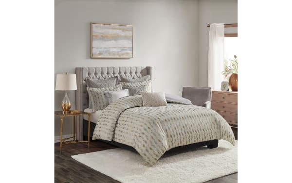Bedroom with a tufted gray upholstered bed featuring a beige and gray patterned comforter set, matching pillows, a gold-accented side table with a glass lamp, a gray armchair, wooden dresser, and abstract wall art.