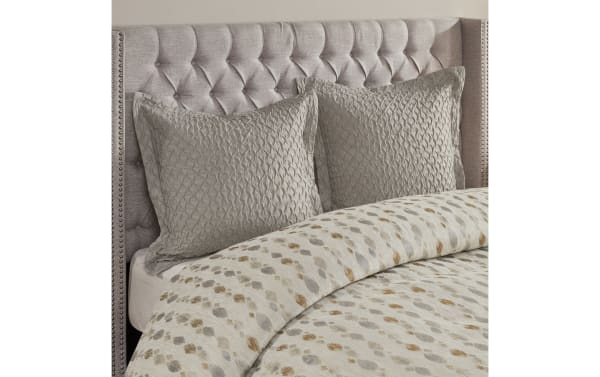 Tufted beige upholstered headboard with silver nailhead trim, paired with textured taupe quilted pillow shams and a patterned beige and gray comforter.