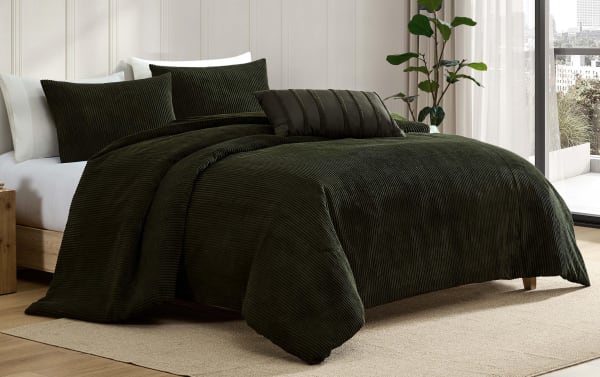 Dark green corduroy textured bedding set on a wooden bed frame, featuring a comforter, multiple pillows, and a decorative cushion in a modern bedroom with natural light.