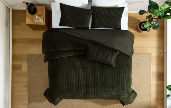 Dark green corduroy textured bedding set with matching pillows and a black accent pillow on a wooden floor with a beige rug and a bedside table.