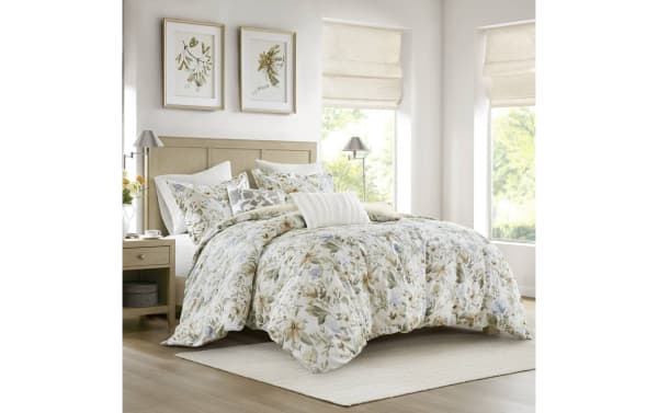 Bed with floral-patterned comforter and matching pillows in beige, green, and light blue tones, set in a bright, modern bedroom with wooden furniture and framed botanical prints.