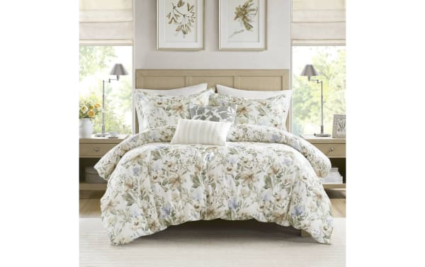 Bed with beige wooden headboard dressed in floral-patterned comforter and pillow set in soft beige, green, and blue tones, flanked by matching nightstands and lamps.