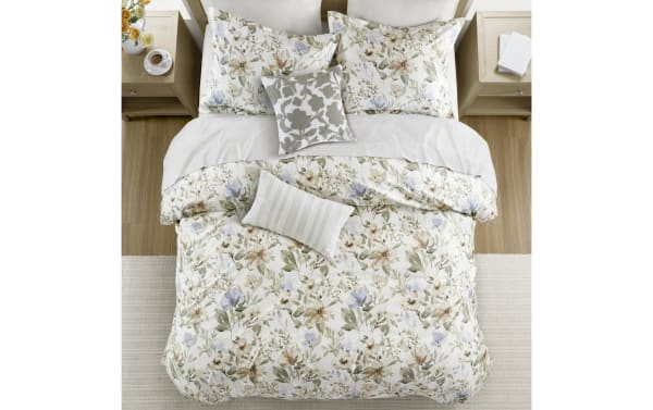 Floral-patterned bedding set with beige, green, and light blue flowers on a white background, featuring matching pillowcases and decorative cushions on a wooden bed.