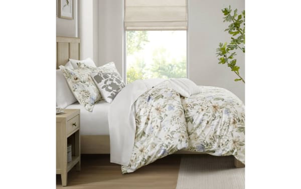 Bed with floral-patterned comforter and matching pillows in soft beige, green, and blue tones, featuring a light wood frame and neutral decor.