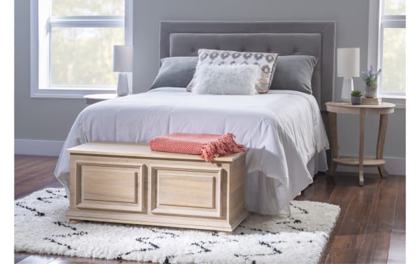 Modern bedroom with a gray upholstered bed, white bedding, decorative pillows, wooden storage bench with a coral throw, round wooden nightstands, table lamps, and a white patterned rug on dark hardwood floor.