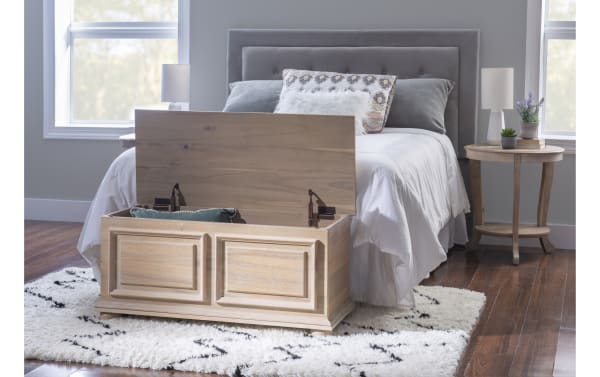 Wooden storage bench with paneled design and lift-up lid at the foot of a gray upholstered bed in a modern bedroom with hardwood floors and white shag rug.