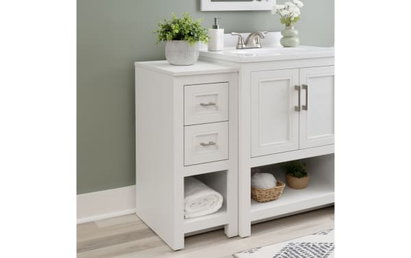 White wooden bathroom vanity side cabinet with two drawers, open shelf with rolled towel, and decorative plants on top, next to sink.