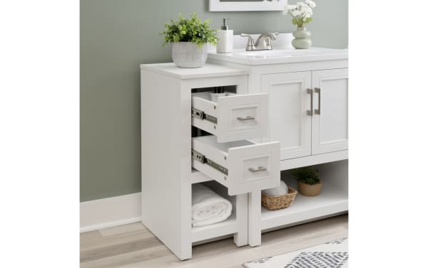 White wooden bathroom storage cabinet with two open drawers, rolled towel on bottom shelf, and decorative plants on top, next to white vanity.