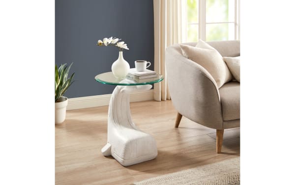 Modern white sculptural side table with a curved hand-shaped base and round glass top, styled with a white vase, books, and coffee cup.