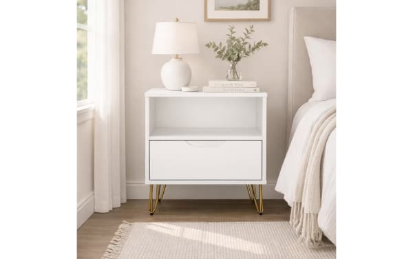 White modern bedside table with gold hairpin legs, single drawer, open shelf, topped with a white lamp, books, and a glass vase with greenery.