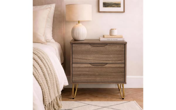 Wooden bedside table with two drawers, gold metal hairpin legs, and a white ceramic lamp with a beige shade on top, next to a bed with cream bedding and a knitted throw.
