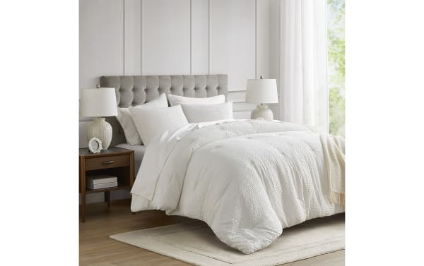 Modern bedroom featuring a queen-size bed with a tufted gray fabric headboard, white textured comforter, matching pillows, wooden nightstands, and ceramic table lamps.