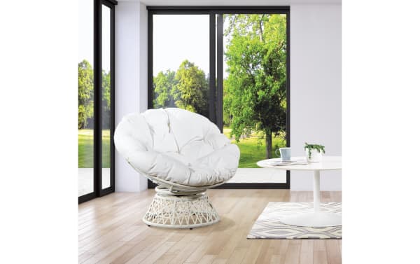 White cushioned papasan chair with a woven rattan base in a bright room with large glass windows overlooking green trees, paired with a white round table on a patterned rug.
