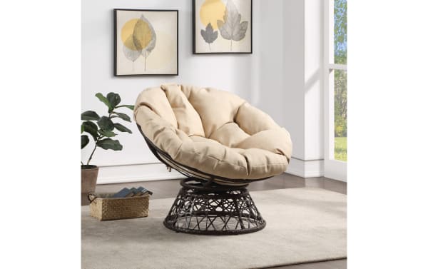 Beige cushioned papasan chair with a black woven rattan frame, placed on a light gray rug in a bright room with framed leaf art and a potted plant.