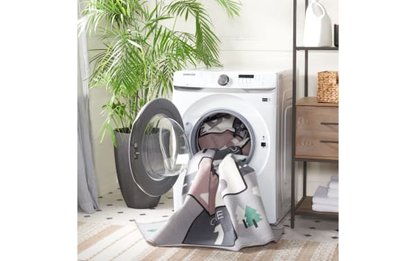 Samsung white front-load washing machine with open door and patterned blanket partially inside, set in a bright laundry room with plant and wooden shelving.