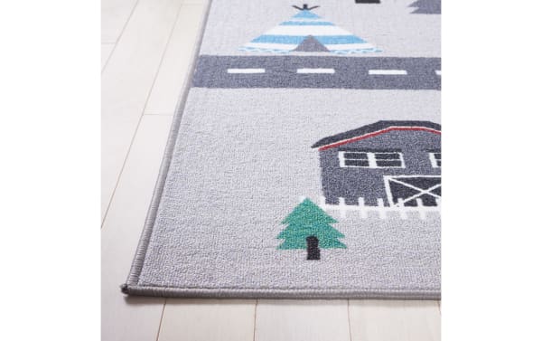 Children's play rug with a gray background featuring a blue and white teepee, a dark gray barn with red trim, a green pine tree, and a road. The rug has a soft, textured surface and a stitched edge, suitable for playrooms or kids' bedrooms.