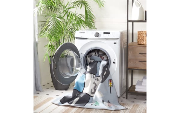 Front-loading Samsung washing machine with an open door, featuring a light blue, gray, and white Happy Hopper 2' x 8' transitional fabric runner with a playful road and number block pattern partially inside. The synthetic polyamide rug is washable, slip-resistant, and OEKO-TEX certified, ideal for safe, easy-clean children's play areas.