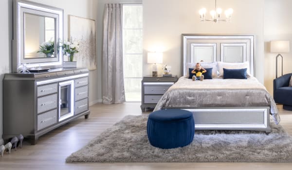Luxurious bedroom with a silver glitter bed frame, matching nightstand and dresser, plush gray bedding, blue velvet ottoman, and soft shag rug.