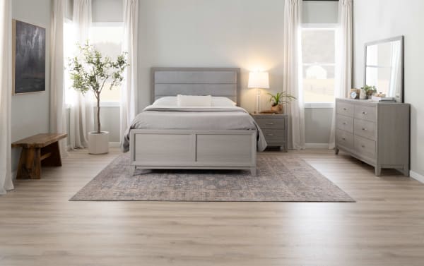 Modern bedroom with light gray wooden bed frame, matching nightstand and dresser, soft gray bedding, large area rug, natural wood bench, potted plant, and neutral decor.