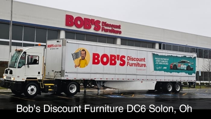 White semi-truck and trailer with large red text "BOB'S Discount Furniture" and promotional graphics, parked in front of a BOB'S Discount Furniture store.