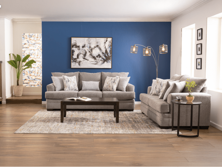 A cozy living room features a gray sofa set with patterned pillows, a dark wood coffee table, and a modern floor lamp against a blue accent wall and abstract art.