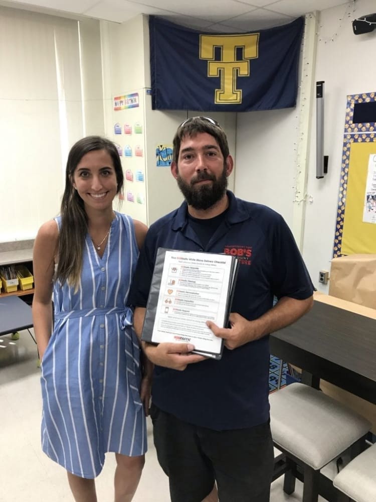 Two people standing in a classroom with a blue and yellow Texas Tech University flag on the wall. One person wears a blue and white striped sleeveless dress with buttons and a waist tie. The other wears a navy blue polo shirt with red text reading "Bob's Discount Furniture," holding a laminated checklist titled "Your 100% White Glove Delivery Checklist" with sections on Arrival, Protection, Preparation, Assembly, and Clean Up.