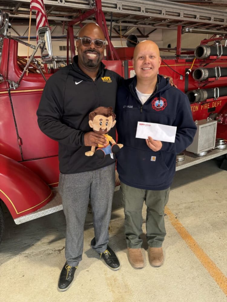 Two men standing in front of a red fire truck, one wearing a black jacket with "BOB'S" logo holding a plush toy, the other in a navy blue firefighter hoodie labeled "LT VALLE" holding an envelope with "BOB'S" printed on it.
