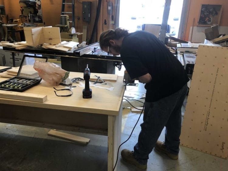 Person using a jigsaw to cut wood on a workbench in a woodworking shop with tools, safety glasses, and wood pieces nearby.