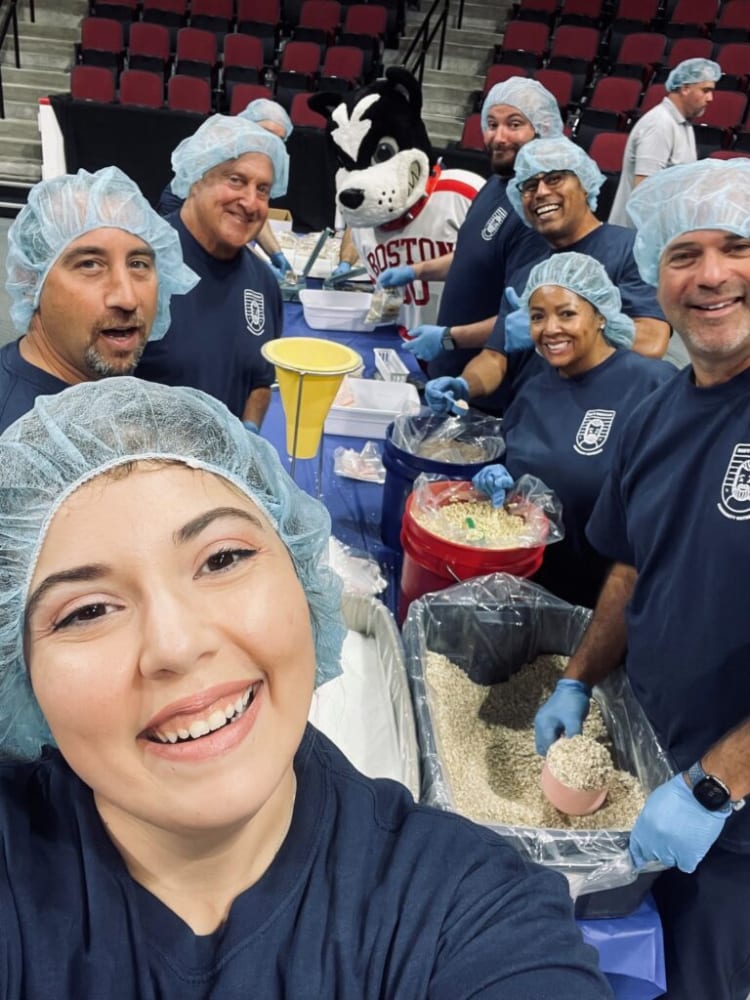 Volunteers wearing blue hairnets and gloves packing dry grains into plastic bags at a community food packaging event in an indoor arena.