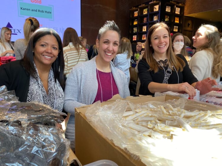 Three women standing behind tables filled with packaged items, including a large box of individually wrapped white and yellow sticks, at an indoor event with a screen displaying "Karen and Rob Hale" in the background.